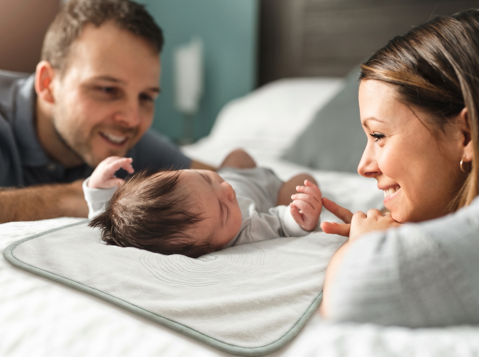 Two parents lying on a bed on either side of their newborn, who lies on the lillypad — a cream plush pad with sage trim and pond-ripple quilting