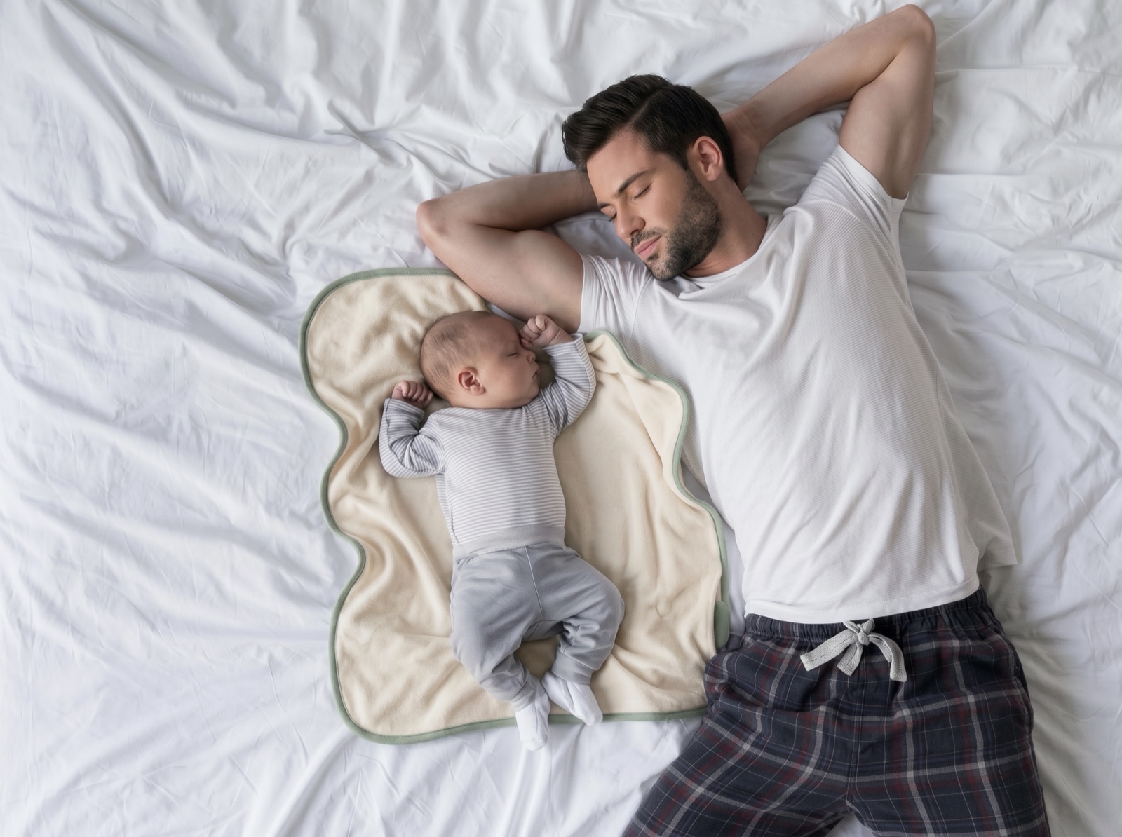 Top-down view of a father dozing on white bedding beside his newborn, who sleeps on the lillypad — a cream plush pad with sage binding