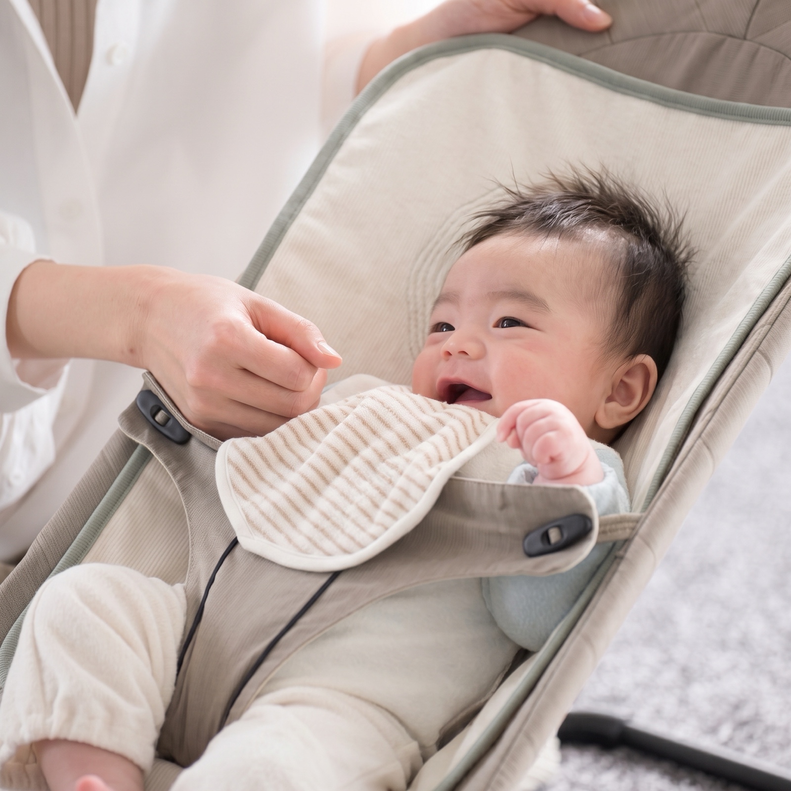 A smiling baby lying in a bouncer draped with the lillypad while a parent leans in close