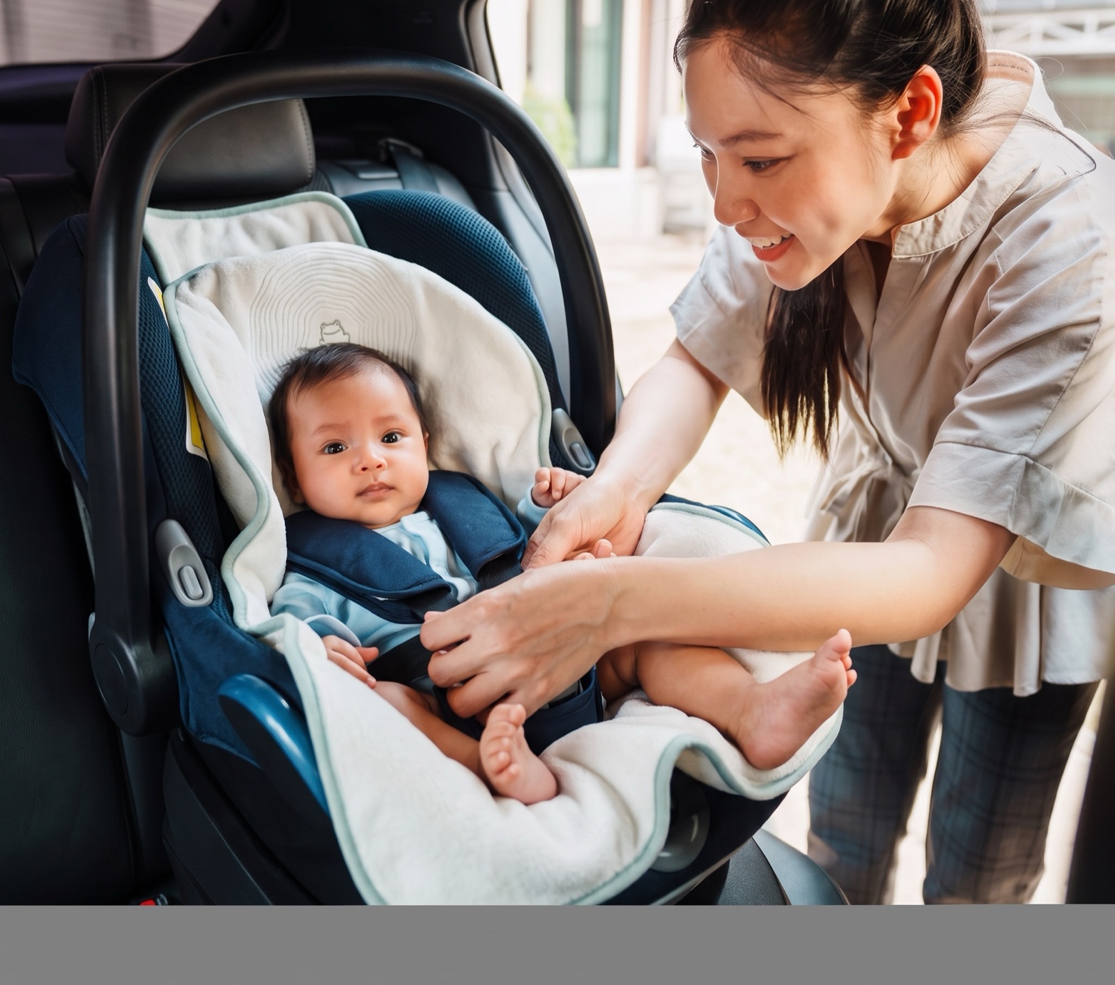 A mother buckling her bright-eyed baby into a carseat lined with the lillypad, the frog and ripple quilting visible behind the baby's head