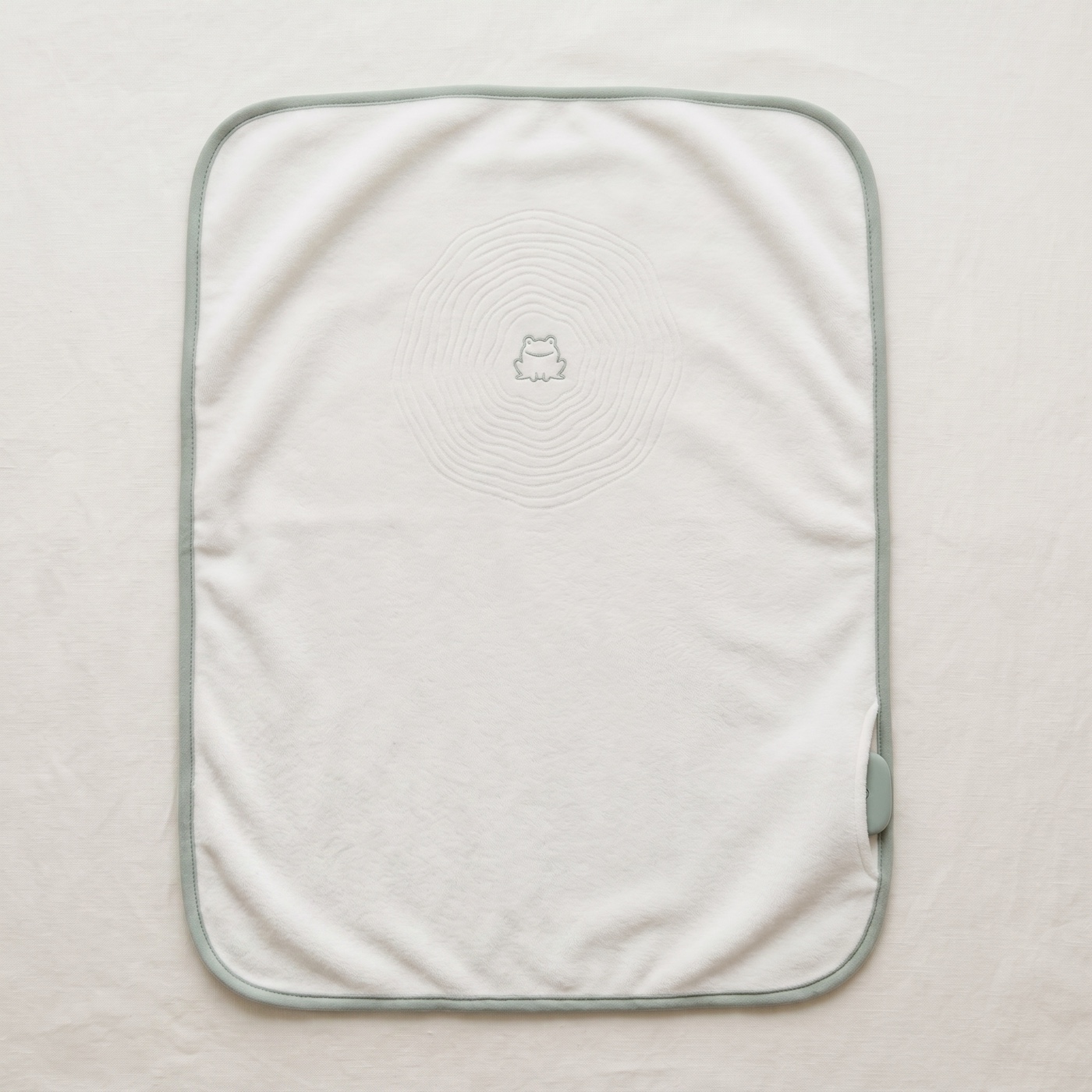 The lillypad flat on linen: cream plush, sage binding, pond-ripple quilting with a small embroidered frog at the center